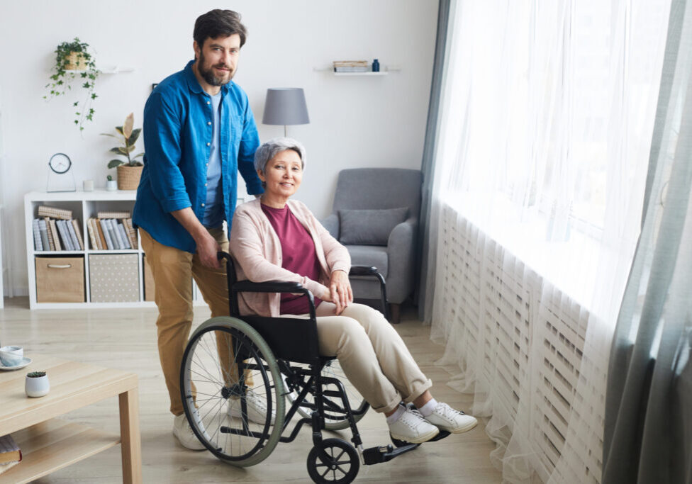 Disabled senior woman sitting in wheelchair and smiling at camera with man standing behind her they are in the room at home